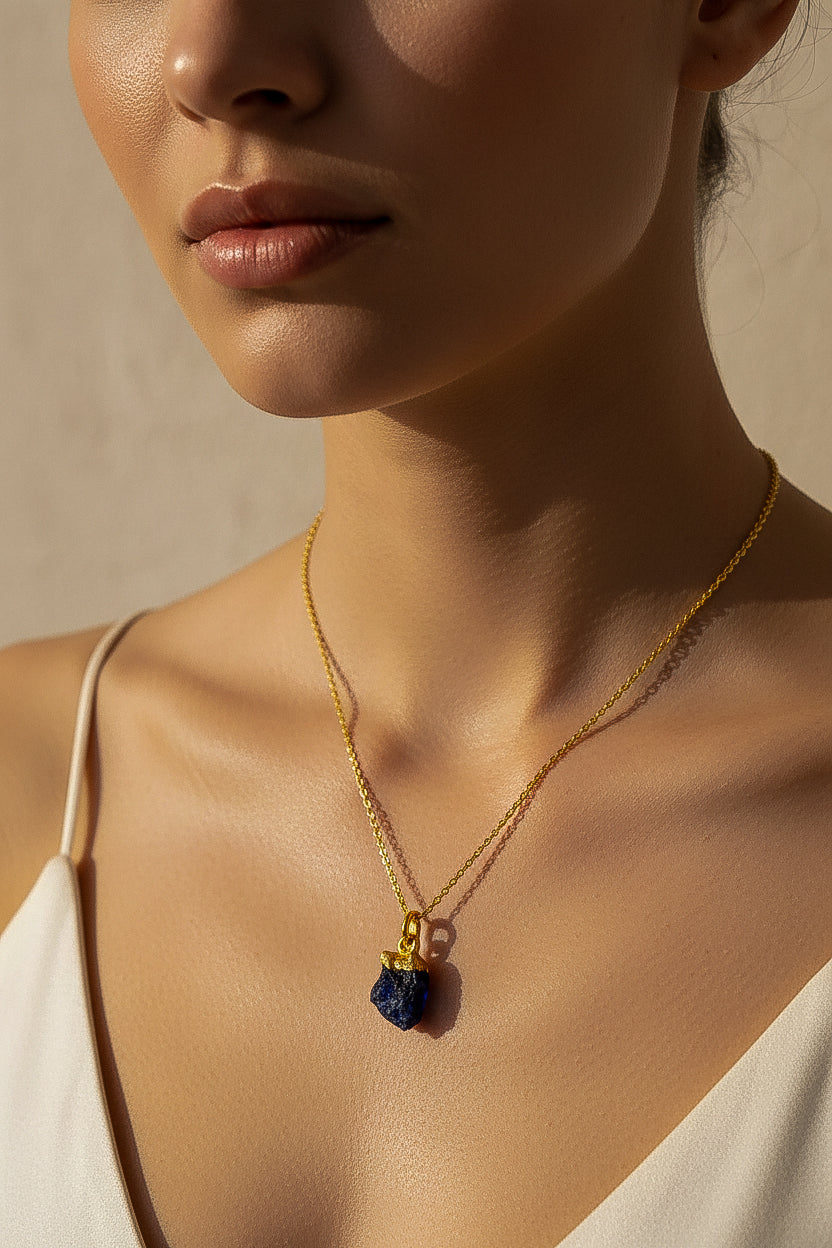Close-up of a woman wearing a gold necklace with a dark blue Raw Sapphire crystal pendant against a neutral background