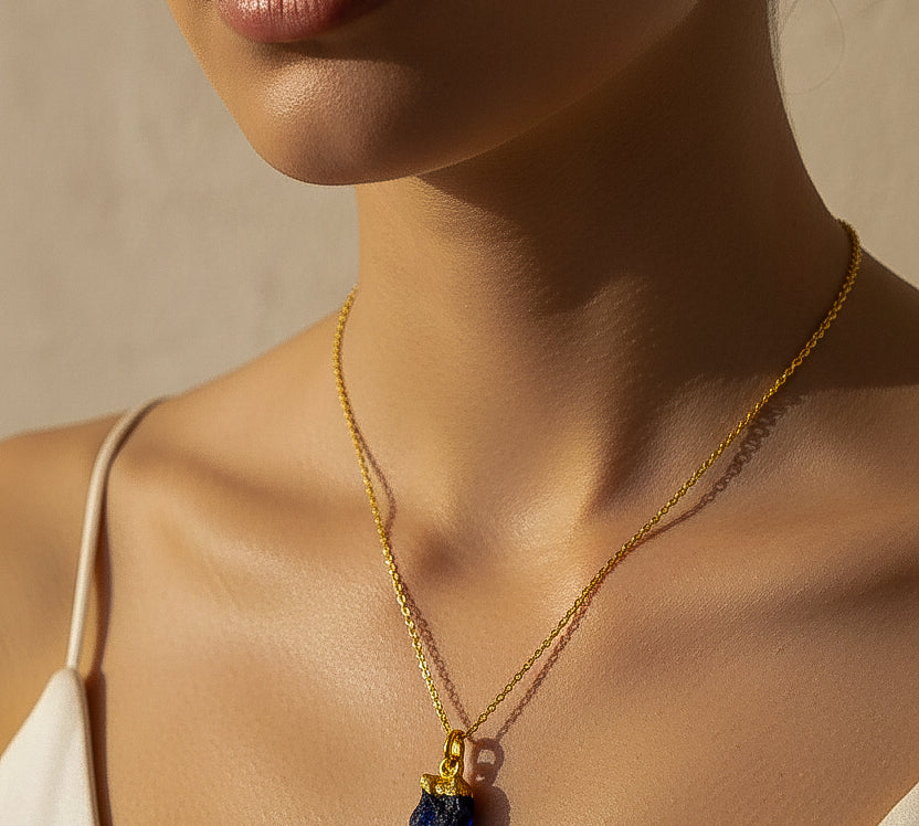 Close-up of a woman wearing a gold necklace with a dark blue Raw Sapphire crystal pendant against a neutral background