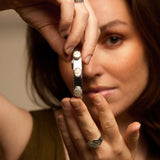 Woman holding a silver cuff with shell gemstones up to her face, wearing a silver shell ring, with a neutral background